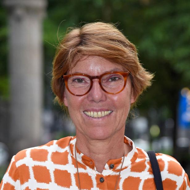 Bettina Böttinger lächelt mit kurzen roten Haaren und oranger Brille. Sie trägt eine orange-weiß gemusterte Bluse.