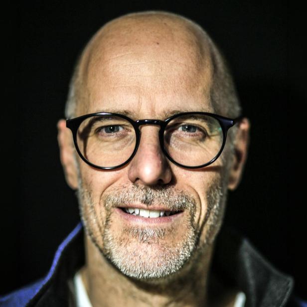 Nahaufnahme des lächelnden Patrick Schubert mit schütterem Haarwuchs, Brille und Bart vor dunklem Hintergrund.