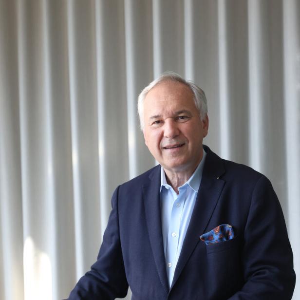 Walter Rosenkranz lehnt an einem Tisch vor hellen Vorhängen und lächelt.