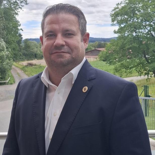 Ein Mann im dunklen Anzug und weißem Hemd steht im Freien vor grüner Landschaft.