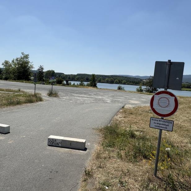 Sperrschilder blockieren eine Straße am Seeufer mit Blick auf Wasser und Hügel.