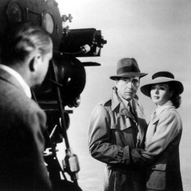 Humphrey Bogart und Ingrid Bergman stehen in Trenchcoats und Hüten vor einer Filmkamera.