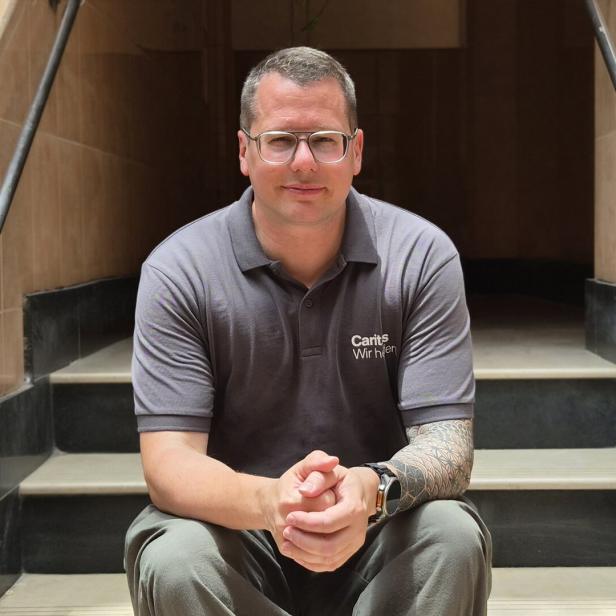 Christoph Riedl mit Brille und Tattoo sitzt auf einer Treppe vor einem dunklen Eingang.