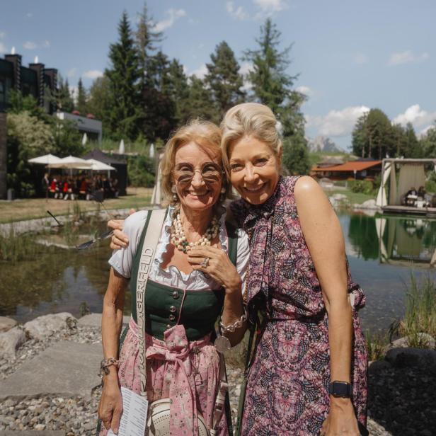 Zwei Frauen lächeln vor einem Naturbadesee mit Bühnenzelt im Alpenresort.