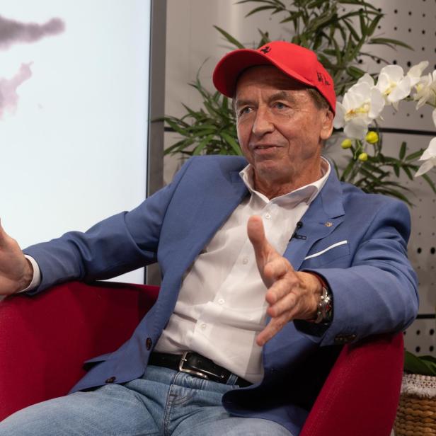 Heribert Kasper sitzt in einem roten Sessel, trägt ein blaues Sakko sowie eine rote Kappe und gestikuliert während eines Gesprächs.
