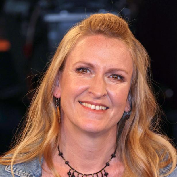Eine lächelnde Frau mit blondem Haar und Jeansjacke vor dunklem Hintergrund.