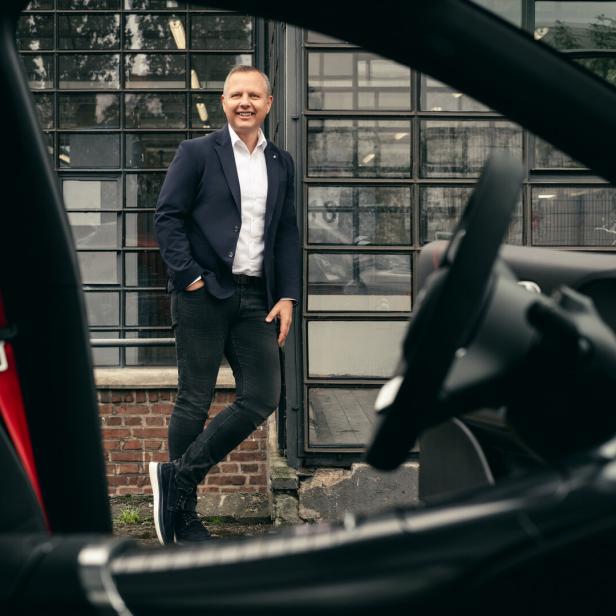 Mann im blauen Blazer steht lächelnd vor einem Auto und einem Industriegebäude.