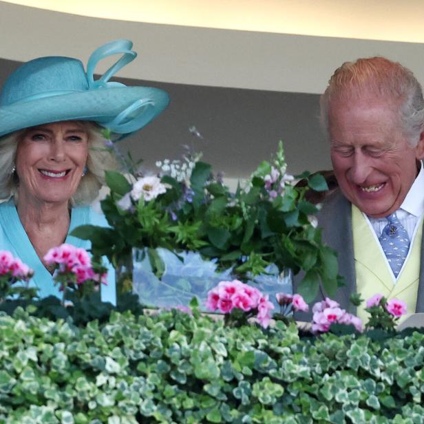 König Charles III. und Königin Camilla lächeln gemeinsam hinter einem mit Blumen geschmückten Balkon.