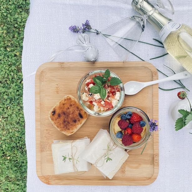 Ein Picknick auf einer Wiese mit einem Holzbrett, das belegte Sandwiches, einen Tomaten-Feta-Salat, ein Beeren-Dessert und Sekt umfasst.