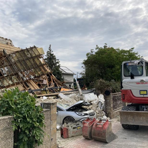 Abrissstelle mit eingestürztem Haus, verschüttetem Auto und Bagger neben Trümmern.