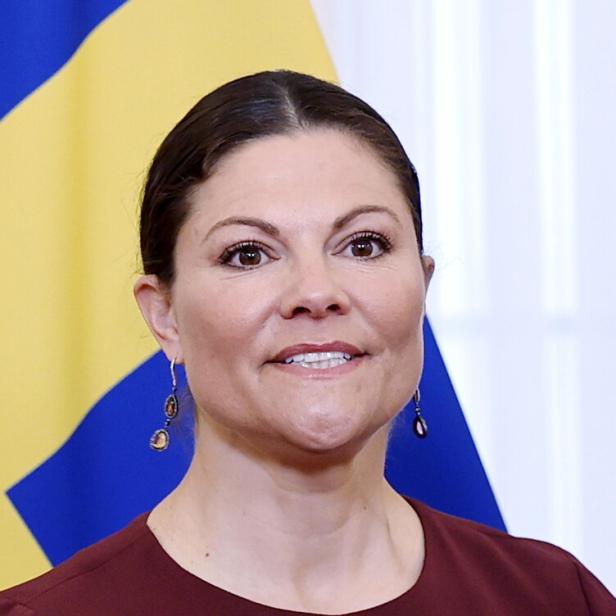 Kronprinzessin Victoria von Schweden trägt ein dunkelrotes Oberteil vor der schwedischen Flagge.