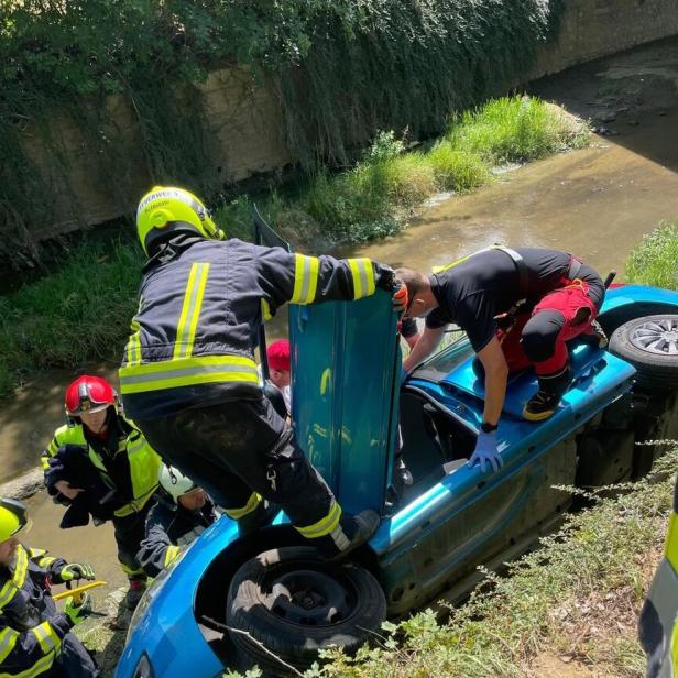 Feuerwehrkräfte retten eine Person aus einem auf der Seite liegenden blauen Auto am Bachufer.