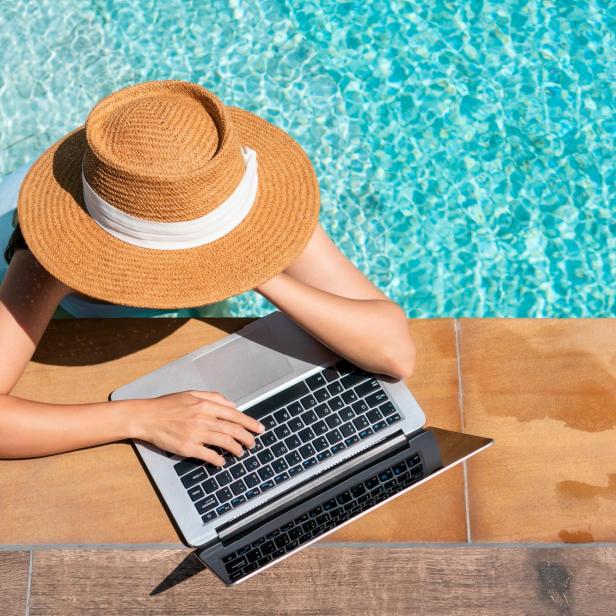 Eine Frau ist in einem Pool und trägt einen Sonnenhut und arbeitet an einem Laptop