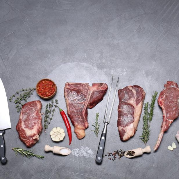 Vier rohe Steaks mit Messern, Kräutern, Knoblauch, Chilischoten und Gewürzen auf grauem Hintergrund.