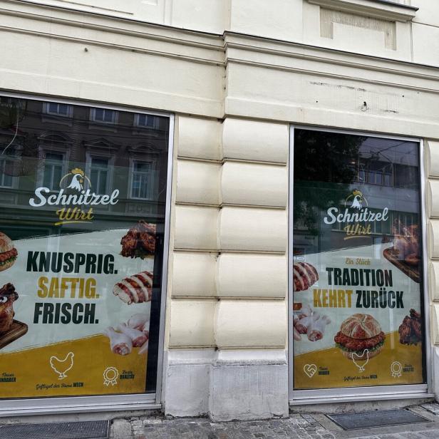 Schaufenster eines Schnitzel-Lokals mit Werbeplakaten für Burger, Hähnchen und Slogans wie „Knusprig. Saftig. Frisch.“.