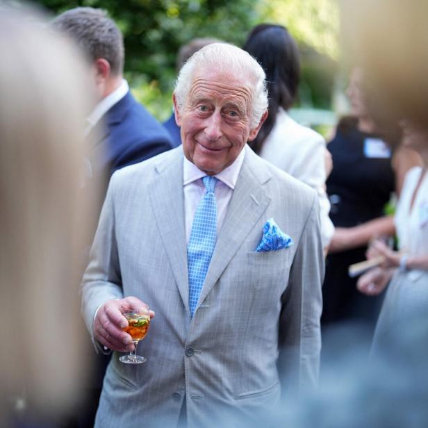 König Charles III. lächelt in einem hellen Anzug mit blauem Einstecktuch und hält ein Glas in der Hand, umgeben von Gästen bei einer Veranstaltung.
