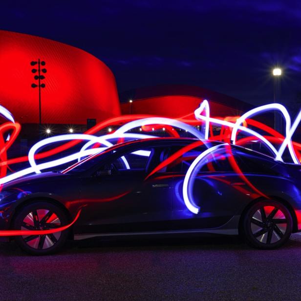 Ein schwarzer Elektro-Pkw steht vor einem rot beleuchteten Gebäude, umgeben von roten und weißen Lichtspuren.