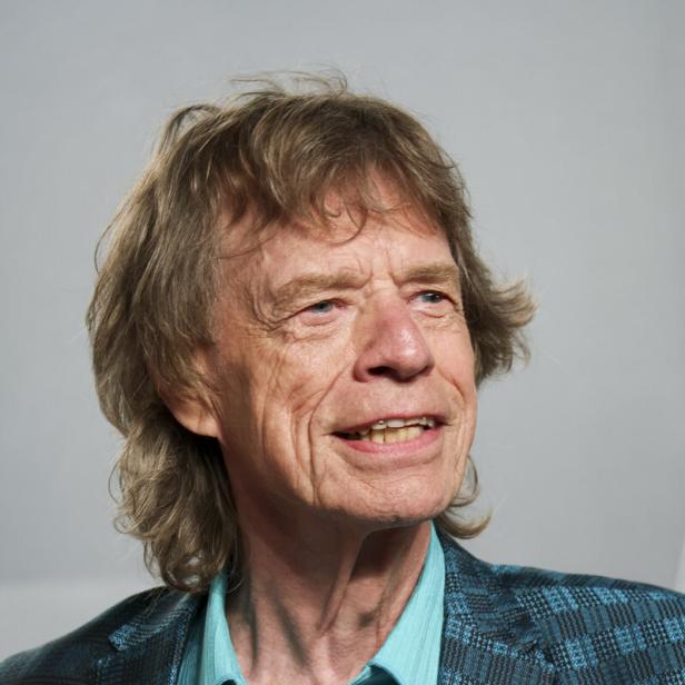 Mick Jagger lächelt vor einem grauen Hintergrund in einem türkisfarbenen Hemd und einem gemusterten Sakko.