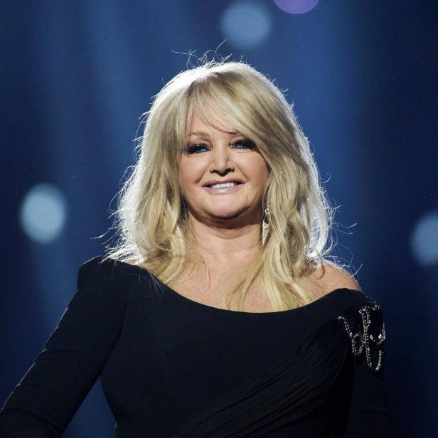 Bonnie Tyler lächelt vor einem dunklen, blau beleuchteten Hintergrund in die Kamera.