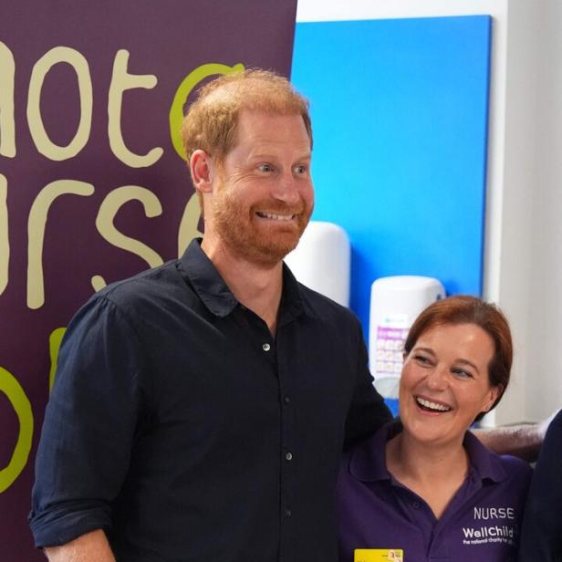 Prinz Harry lächelt neben einer Krankenschwester der Wohltätigkeitsorganisation WellChild vor einem Werbebanner.