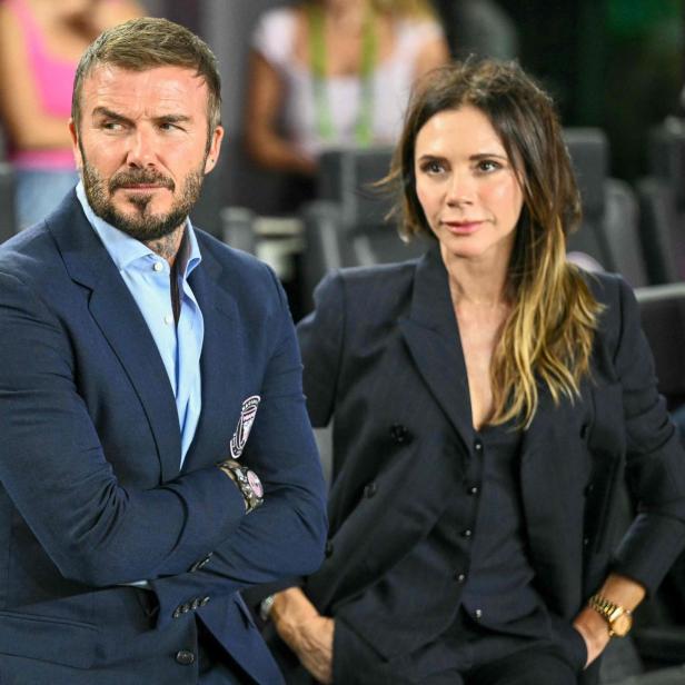 David und Victoria Beckham sitzen nebeneinander in eleganten dunklen Anzügen auf einer Tribüne.