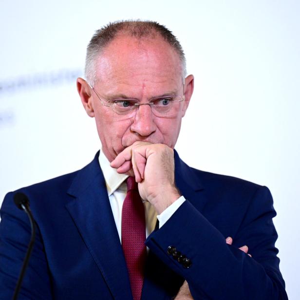Ein Mann im Anzug und mit Brille steht nachdenklich an einem Rednerpult, die Hand vor dem Mund.