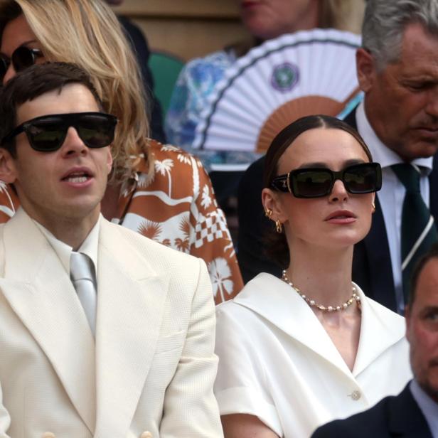 Keira Knightley trägt eine Sonnenbrille und ein weißes Oberteil, während sie neben anderen Zuschauern auf der Tribüne in Wimbledon sitzt.