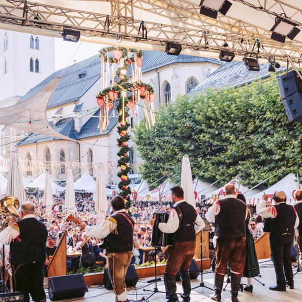 Eine Musikkapelle in Tracht spielt auf einer Bühne vor einem Maibaum und einer Menschenmenge auf einem Marktplatz.