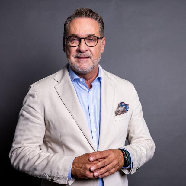 Heinz Christian Strache mit Brille, grauem Bart und hellem Sakko steht vor einem grauen Hintergrund.