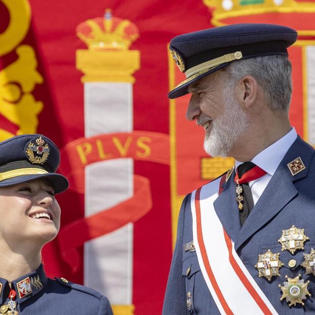 König Felipe VI. von Spanien lächelt seiner Tochter Prinzessin Leonor in Militäruniform vor einer spanischen Flagge zu.