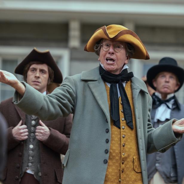 Ein Mann (Larry David) mit Brille und in historischer Kleidung mit Dreispitz gestikuliert lebhaft vor einer Gruppe weiterer Personen in zeitgenössischen Kostümen.