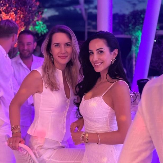 Zwei Frauen in weißen Outfits lächeln bei einer Party im violetten Licht.