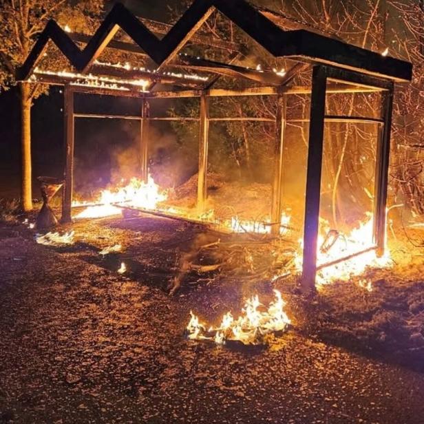 Ein offener Unterstand aus Metall steht in Flammen, mit Feuer auf dem Boden und an den Pfosten.