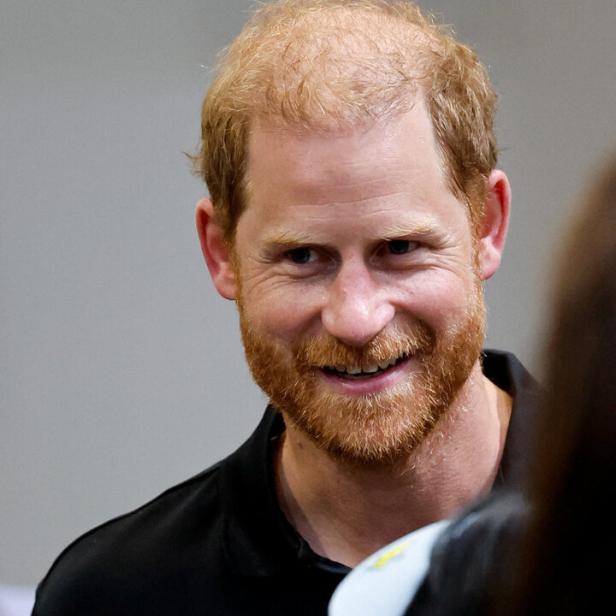 Prinz Harry lächelt freundlich in einem schwarzen Poloshirt vor einem neutralen Hintergrund.