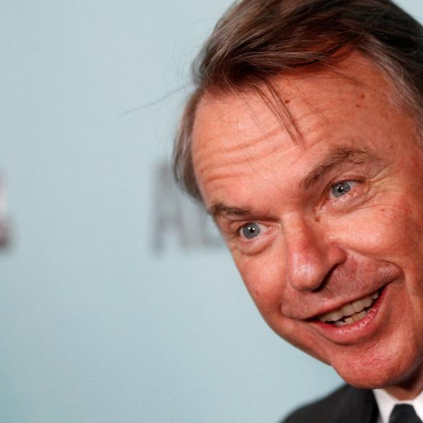 Der Schauspieler Sam Neill lächelt vor einem hellblauen Hintergrund.