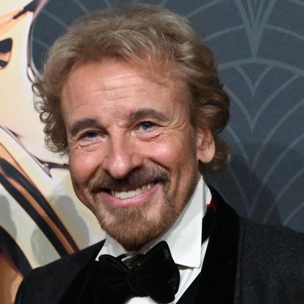 Thomas Gottschalk lächelt in einem dunklen Anzug mit weißem Hemd und schwarzer Fliege vor einem gemusterten Hintergrund.