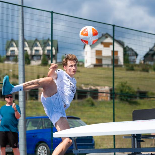 Ein junger Mann in weißer Sportkleidung spielt Teqball auf einem blauen Außenplatz.