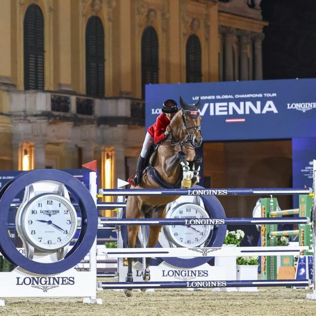 Springreiter überwindet ein Longines-Hindernis bei Nacht vor historischer Fassade.