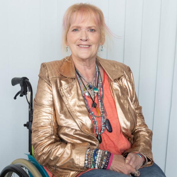 Gaby Köster mit blondem Haar trägt eine goldene Jacke und viele Halsketten, während sie in einem Rollstuhl sitzt.