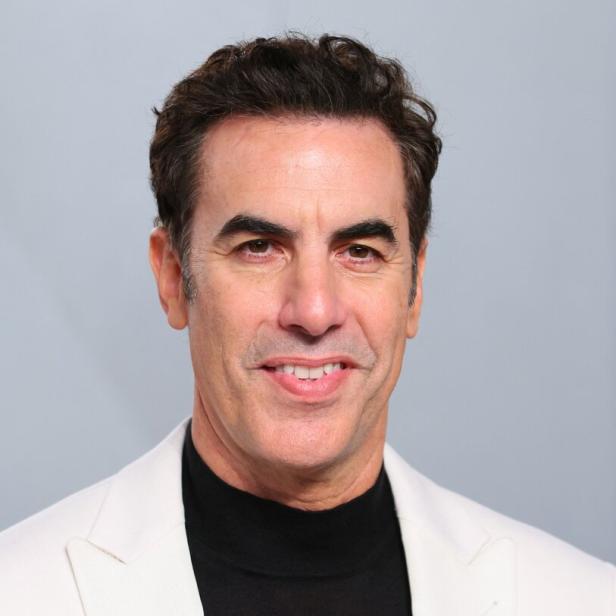 Sacha Baron Cohen lächelt vor einem grauen Hintergrund in einem weißen Sakko über einem schwarzen Rollkragenpullover.