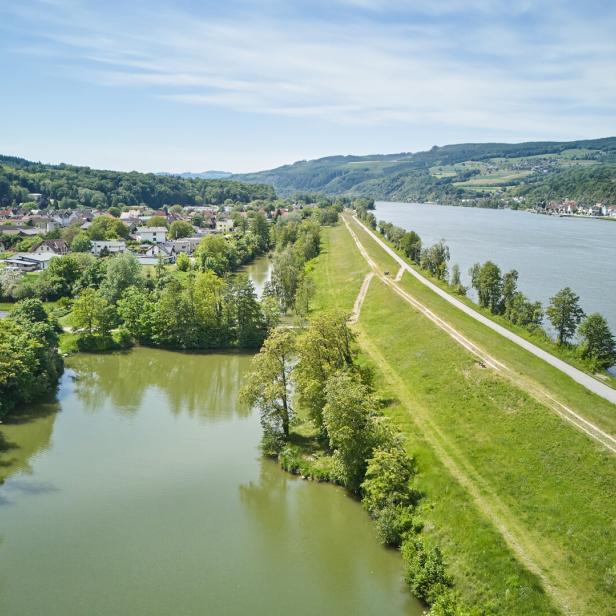 Luftaufnahme eines Flusstals mit Damm, Uferweg, Wohnhäusern und bewaldeten Hügeln.