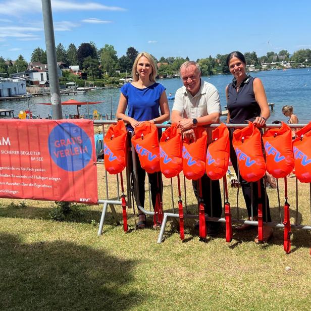 Drei Personen stehen am See vor einer Reihe orangefarbener Schwimmbojen und einem KFV-Banner.