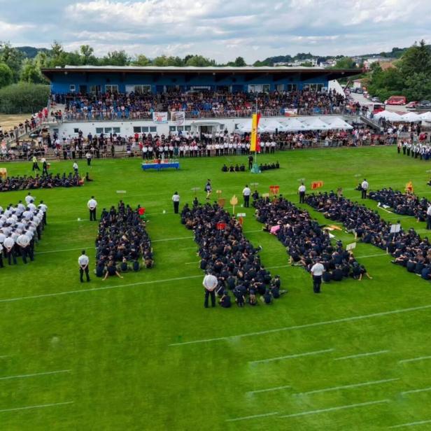Zahlreiche Feuerwehrleute in Uniform sitzen in Gruppen auf einem Sportplatz vor einer voll besetzten Tribüne bei einer feierlichen Veranstaltung.