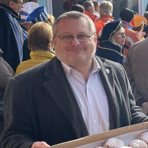 Mann lächelt in die Kamera und hält eine Kiste mit Puderzucker-Krapfen auf einem vollen Festplatz.