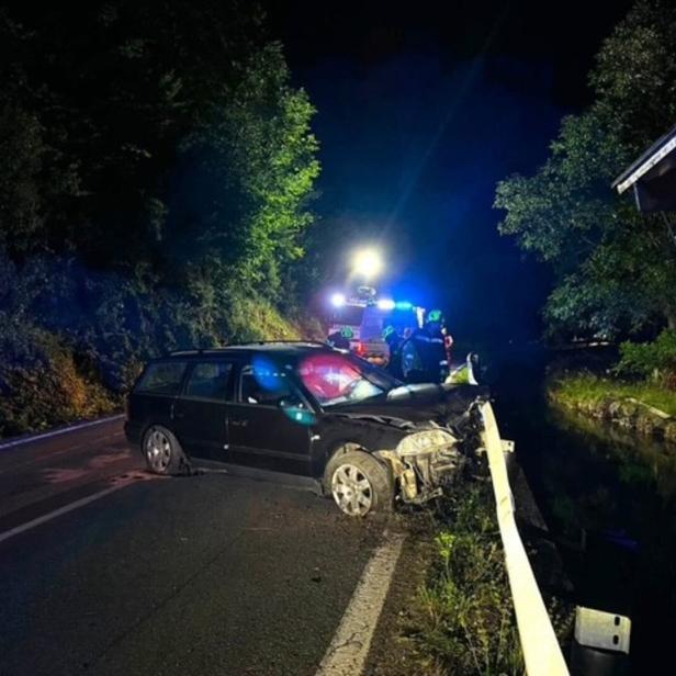 Nächtliche Unfallstelle: Ein schwarzer Kombi mit Frontschaden steht neben der Leitplanke, Rettungskräfte sind vor Ort.