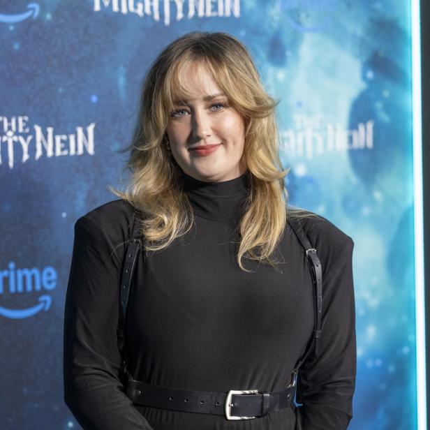 Ashley Johnson lächelt vor einem blauen Hintergrund mit dem Schriftzug „The Mighty Nein“ in die Kamera.