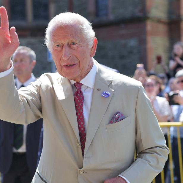 König Charles III. winkt in einem beigen Anzug und mit einer roten Krawatte einer Menschenmenge zu.