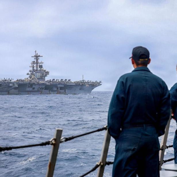 Matrosen blicken von einem Schiff auf einen Flugzeugträger im Meer, während ein Kampfjet startet.