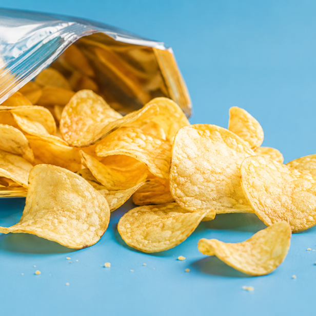 Eine geöffnete Tüte mit Kartoffelchips liegt auf einem hellblauen Untergrund, wobei einige Chips aus der Packung herausgefallen sind.