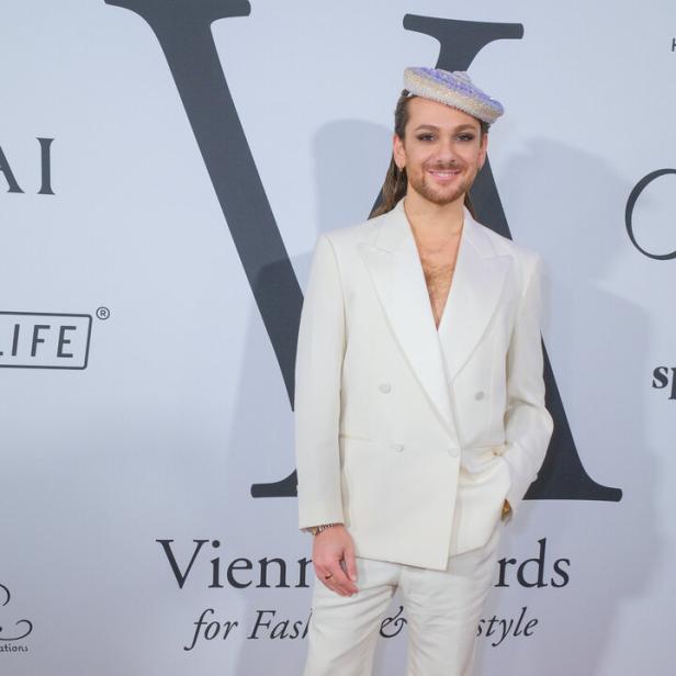 Riccardo Simonetti trägt einen weißen Anzug und eine verzierte Kopfbedeckung vor einer Wand der Vienna Awards for Fashion & Lifestyle.